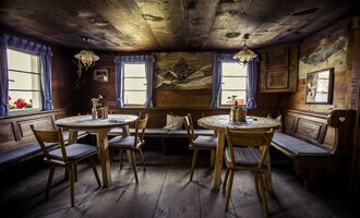 A cozy room with wooden paneling and two round tables. The windows offer a picturesque view and the decor exudes rustic charm. | © Kleinwalsertal Tourismus | Oliver Farys