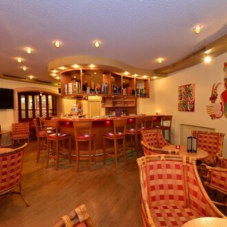 A cozy bar with welcoming chairs and warm lighting. In the background, there is a well-stocked bar and colorful wall art. | © IFA Hotel Alpenhof Wildental
