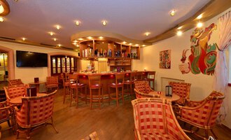 A cozy bar with welcoming chairs and warm lighting. In the background, there is a well-stocked bar and colorful wall art. | © IFA Hotel Alpenhof Wildental