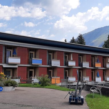 A modern building with several balconies in a green environment. In the background, mountains and a blue sky can be seen. | © Kleinwalsertal Tourismus | Veronika Senn