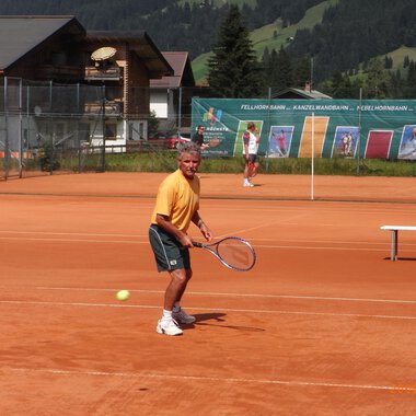 A man is playing tennis on a red court with a view of the mountains. In the background, other players and a tennis wall can be seen. | © Sportverein Casino Kleinwalsertal