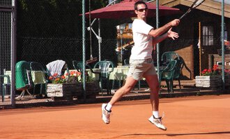 A man is playing tennis on a sand court. In the background, there are chairs and tables as well as a few plants. | © Sportverein Casino Kleinwalsertal