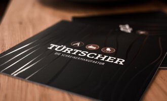 Ein elegantes Menüheft mit der Aufschrift „TÜRTSCHER – die Schreiner-Manufaktur“. Es zeigt verschiedene Holzsymbole auf einem dunklen Hintergrund. | © Türtscher Die Schreinermanufaktur