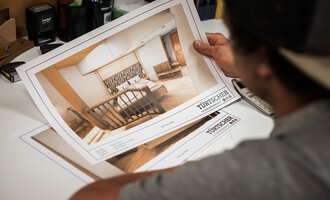 Eine Person betrachtet Pläne für ein Innenraumdesign. Die Pläne zeigen ein modernes, einladendes Zimmer mit einem Bett und einer bequemen Sitzecke. | © Türtscher Die Schreinermanufaktur