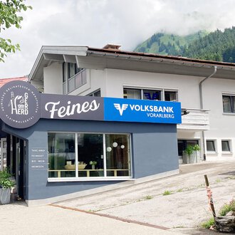 A modern building with a business sign "Feines" and the Volksbank Vorarlberg. In the background, green hills and a clear sky can be seen. | © Volksbank Vorarlberg e. Gen.