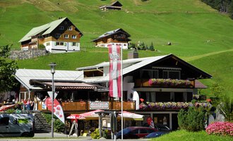 A beautiful alpine building with a terrace and colorful flowers. In the background, green meadows and more traditional houses can be seen. | © Cafe Baad Grund | Roman Schuster