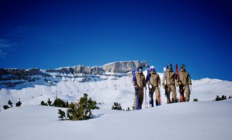 A group of skiers stands on a snow-covered area. In the background, you can see impressive mountain peaks and a clear blue sky. | © Walser Skischule | Herbert Jochum