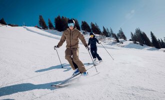 Two skiers are skiing down the slope. The sky is clear and the surroundings are covered in snow. | © Walser Skischule | Ole Zumpolle