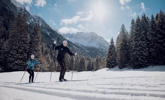 Two people cross-country skiing in a snowy landscape. In the background, majestic mountains and a clear, sunny sky can be seen. | © Walser Skischule | Ole Zumpolle