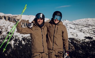 Two skiers are happily posing in the mountains. They are wearing ski clothing and standing in front of a snow-covered landscape. | © Walser Skischule | Ole Zumpolle