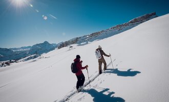 Two hikers are walking through deep snow in the mountains. The sun is shining clearly in the blue sky. | © Walser Skischule | Ole Zumpolle