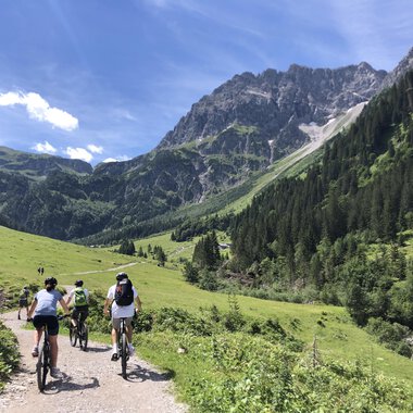 A group of cyclists is riding through a picturesque mountain landscape. The green meadows and the majestic mountains create a beautiful atmosphere. | © WalserBike Tours | Christian Gutermuth