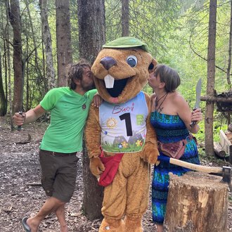 A funny mascot in the shape of a beaver stands between two people in the forest. The humans seem to be having fun and are kissing the mascot on the cheeks. | © Wildnisschule Kleinwalsertal