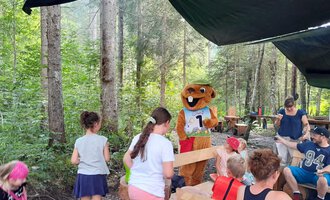 A group of children is standing in a forest and sees a mascot holding a competition about a number. The cheerful atmosphere is enhanced by the green surroundings. | © Wildnisschule Kleinwalsertal