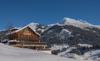 A beautiful chalet in the snow with an impressive mountain in the background. The sky is clear and blue, which makes the winter landscape even more picturesque. | © Wirtshaus Hoheneck | Klaus Ruland