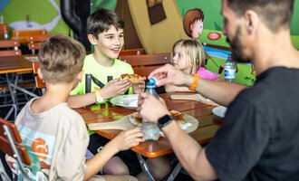 A boy joyfully eats pizza while sitting at a table with two other children. Colorful murals can be seen in the background. | © Wonniland Sonthofen | interSPA Gruppe
