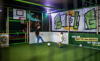 A playing father and his child in an exciting indoor sports arena. They are kicking a soccer ball on green artificial turf under colorful graphics. | © Wonniland Sonthofen | interSPA Gruppe