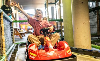 A cheerful boy is riding in a red Bobby-Car and waving excitedly. In the background, other playground equipment can be seen. | © Wonniland Sonthofen | interSPA Gruppe