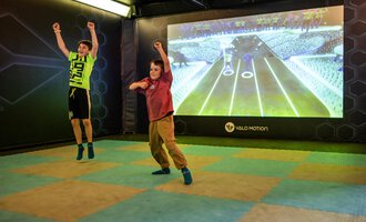 Two children are happily jumping in a playroom. In the background, a large projection screen with an interactive game is visible. | © Wonniland Sonthofen | interSPA Gruppe