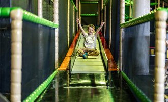 A happy child is sitting on a slide in a playground. The surroundings are colorful and inviting for children. | © Wonniland Sonthofen | interSPA Gruppe