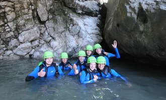 A group of eight people in wetsuits and green helmets is standing in the water between large rocks. They are smiling and giving a thumbs up. | © Rocksolid Canyoning Tours