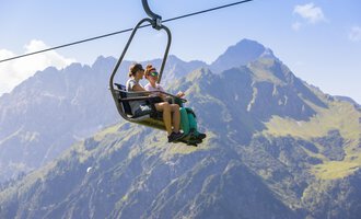 Two people are sitting in a gondola on their way over the mountains. The landscape in the background is majestic with green meadows and high peaks. | © Kleinwalsertal Tourismus | Frank Drechsel