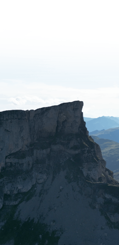 A dramatic mountain peak rising sharply against a pale sky, with distant hills visible in the background.