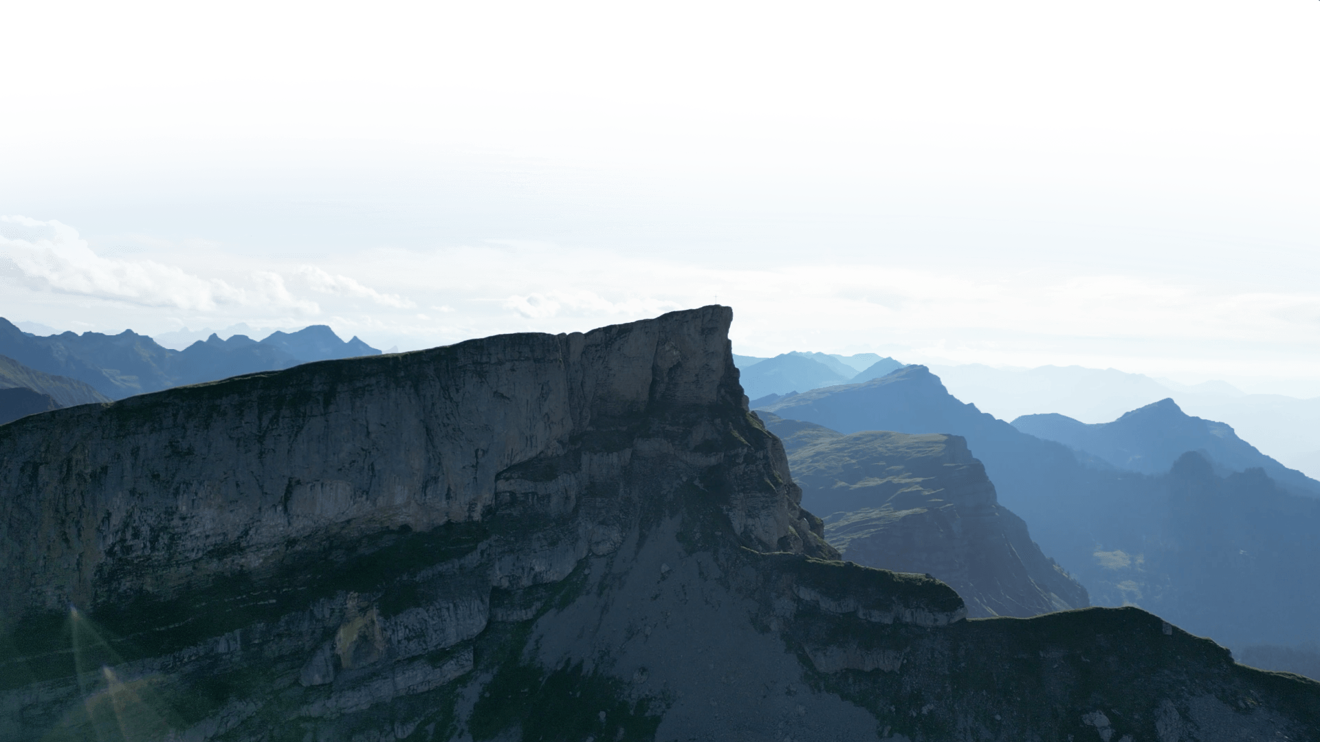 A dramatic mountain skyline with a prominent cliff edge and layered peaks in the background under a cloudy sky.